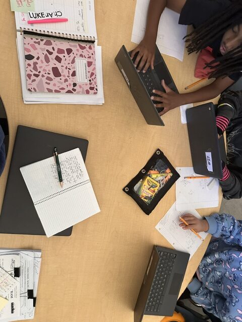 Students working collaboratively at a shared classroom table with notebooks and laptops